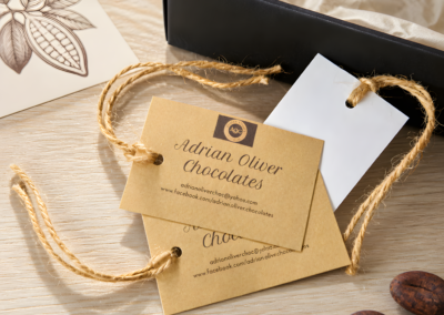 Two craft paper gift tags tied with twine strings showing contact details for "Adrian Oliver Chocolates" placed on a light wood surface. A white card with a hole punch, an open black gift box lined with tissue paper, an botanical cocoa pod illustration, and two cocoa beans rest nearby.