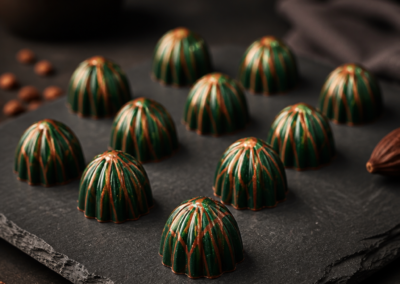 A collection of artisan dark chocolate truffles, featuring a dark green dome with intricate copper and orange hand-painted accents, arranged in rows on a black slate serving platter. The background is a warm, softly blurred setting with a bowl of cocoa beans and a flickering candlelight.