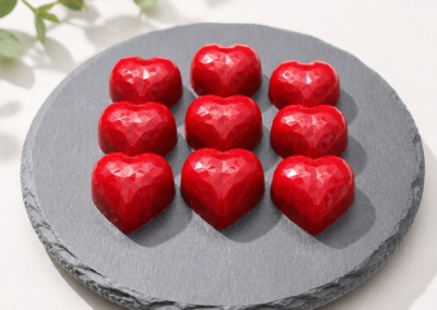 glossy, red heart-shaped ruby chocolates with a faceted geometric pattern, arranged in a 3x3 grid on a round slate platter.